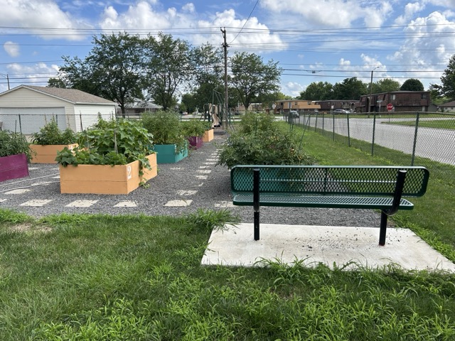 Bourbonnais park shows flower boxes, slides, and a bench. The park is surrounded by a chain link fence.
