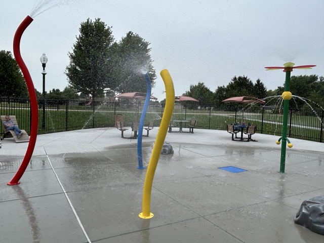 Splashpad surrounded by a black fence. Fun splash pad features are red, blue, and yellow.