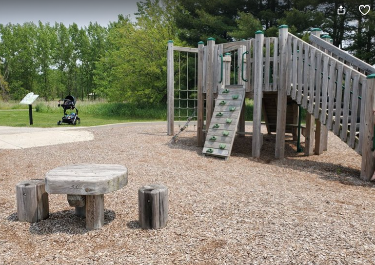 Wooden playground equipment and wooden table and stools in the foreground at Willowhaven Park in Kankakee County.